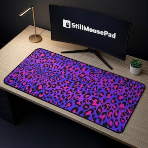 May include: A rectangular mousepad with a vibrant leopard print pattern in shades of purple, pink, and black. The mousepad is placed on a wooden desk, with a computer monitor displaying the text "StillMousePad".