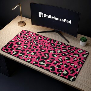 May include: A rectangular desk mousepad featuring a pink leopard print design. The mousepad has black edges and a black, green, and pink pattern. The computer monitor in the background displays the text "StillMousePad".