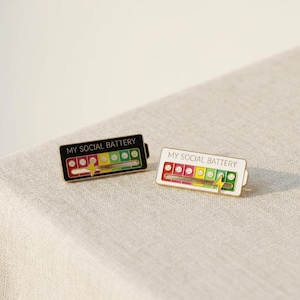 May include: Two enamel pins with the text "MY SOCIAL BATTERY". One pin is black with a yellow, green, and red battery indicator. The other pin is white with a similar indicator. The pins have a lightning bolt design.