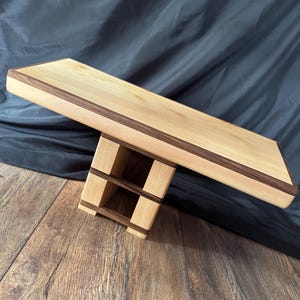May include: A rectangular wooden display stand featuring a light-coloured top surface with a dark wood border. The base is constructed from light wood blocks separated by thin dark wood horizontal strips, creating a tiered, open-frame support structure.