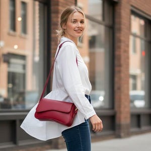 May include: A woman walking on a city sidewalk wearing a white blouse, blue jeans, and white sneakers. A red crossbody bag with a flap closure is worn over the shoulder. The background includes a brick building and large windows.