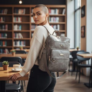 Könnte beinhalten: Ein grauer Kunstleder-Rucksack mit mehreren Fächern und goldfarbener Hardware. Der Rucksack hat einen oberen Griff und verstellbare Schultergurte. Die Tasche wird über der Schulter getragen. Der Hintergrund zeigt eine Bibliothek mit Bücherregalen und Tischen.