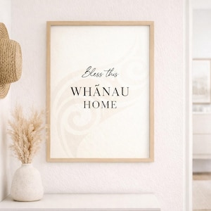 May include: A framed print with the text "Bless this WHĀNAU HOME" in a light wood frame hangs on a white wall. A straw hat hangs on a wall hook to the left of the print. A vase with dried flowers sits on a white shelf below.
