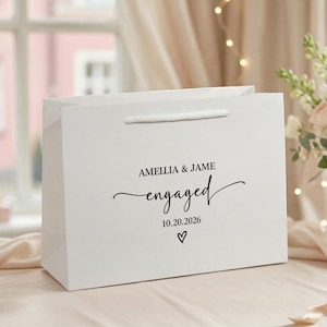 以下が含まれることがあります： AMELLIA & JAME、engaged、10.20.2026という黒い文字と、日付の下に小さなハートのシンボルが印刷された白いギフトバッグ。バッグには白いコードハンドルが付いており、前面は滑らかなマット仕上げになっています。