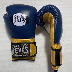 K&ouml;nnte beinhalten: Ein Paar blau-goldene Boxhandschuhe mit dem Cleto Reyes Logo und dem Schriftzug Hecho en Mexico auf dem Handgelenkriemen und dem Handr&uuml;cken. Die Handschuhe haben ein professionelles Design mit einem Klettverschluss und einem 14oz Etikett.