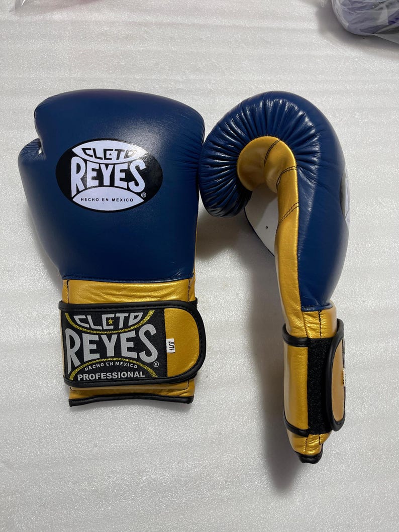 K&ouml;nnte beinhalten: Ein Paar blau-goldene Boxhandschuhe mit dem Cleto Reyes Logo und dem Schriftzug Hecho en Mexico auf dem Handgelenkriemen und dem Handr&uuml;cken. Die Handschuhe haben ein professionelles Design mit einem Klettverschluss und einem 14oz Etikett.