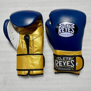 K&ouml;nnte beinhalten: Ein Paar blaue und goldene Boxhandschuhe mit dem Cleto Reyes Logo auf dem Handgelenkriemen und dem Handr&uuml;cken. Die Handschuhe haben eine wei&szlig;e Innenpolsterung und schwarze Details. Der Text Cleto Reyes Hecho en Mexico ist sichtbar.