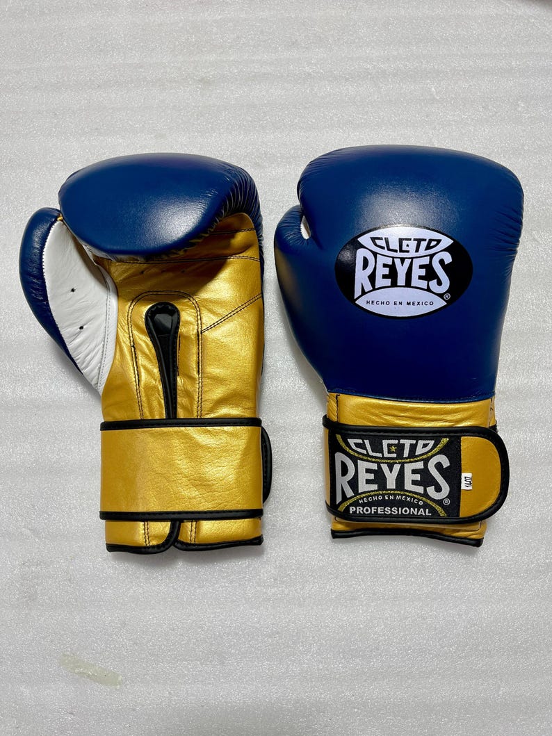 K&ouml;nnte beinhalten: Ein Paar blaue und goldene Boxhandschuhe mit dem Cleto Reyes Logo auf dem Handgelenkriemen und dem Handr&uuml;cken. Die Handschuhe haben eine wei&szlig;e Innenpolsterung und schwarze Details. Der Text Cleto Reyes Hecho en Mexico ist sichtbar.