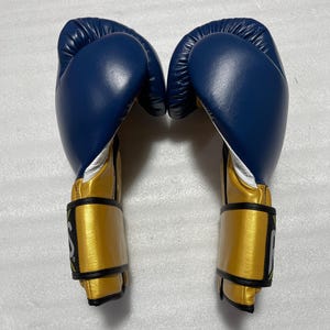 K&ouml;nnte beinhalten: Ein Paar Boxhandschuhe mit einem tiefblauen Oberteil und einer metallisch goldenen Manschette. Die Handschuhe haben ein wei&szlig;es Innenfutter und schwarze R&auml;nder an den Handgelenksriemen, wobei ein teilweises wei&szlig;es Logo an der Seite sichtbar ist.