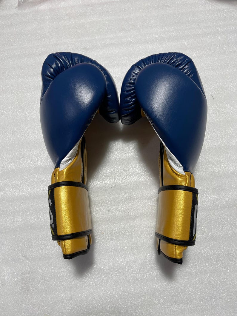 K&ouml;nnte beinhalten: Ein Paar Boxhandschuhe mit einem tiefblauen Oberteil und einer metallisch goldenen Manschette. Die Handschuhe haben ein wei&szlig;es Innenfutter und schwarze R&auml;nder an den Handgelenksriemen, wobei ein teilweises wei&szlig;es Logo an der Seite sichtbar ist.