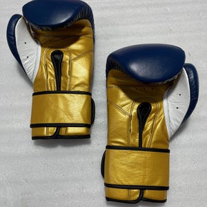 K&ouml;nnte beinhalten: Ein Paar Boxhandschuhe mit metallisch goldener Oberfl&auml;che am Hauptteil und am Handgelenkriemen, kontrastiert durch einen tief dunkelblauen oberen Bereich und eine wei&szlig;e Handfl&auml;che. Die Handschuhe verf&uuml;gen &uuml;ber schwarze Paspeln an den N&auml;hten.