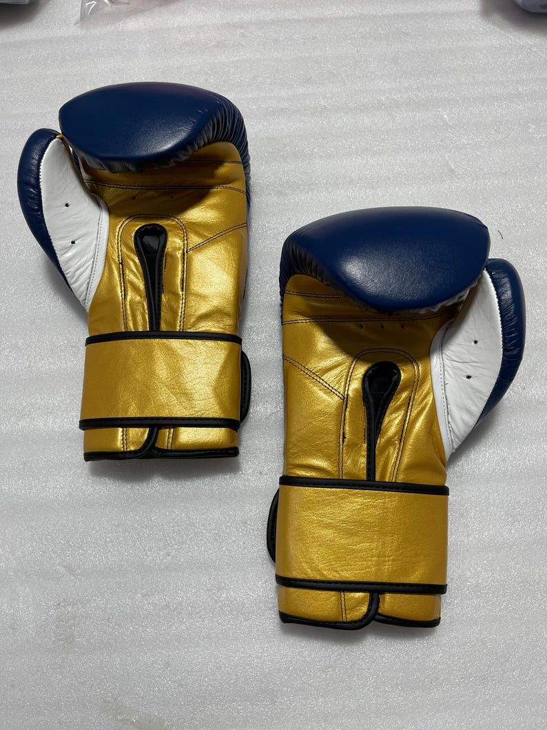 K&ouml;nnte beinhalten: Ein Paar Boxhandschuhe mit metallisch goldener Oberfl&auml;che am Hauptteil und am Handgelenkriemen, kontrastiert durch einen tief dunkelblauen oberen Bereich und eine wei&szlig;e Handfl&auml;che. Die Handschuhe verf&uuml;gen &uuml;ber schwarze Paspeln an den N&auml;hten.