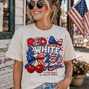 Puede incluir: Camiseta color crema con un diseño patriótico. El gráfico presenta el texto "Red & White 250 America" con lazos y cerezas. La camiseta celebra 250 años de libertad.