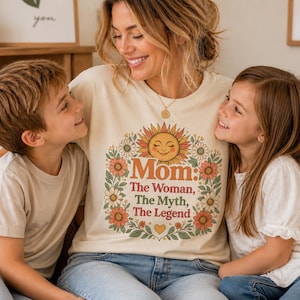 Puede incluir: Una camiseta color crema con el texto "Mom: The Woman, The Myth, The Legend" en naranja y marrón, rodeada por una corona floral. Una mujer y dos niños sonríen y se abrazan.