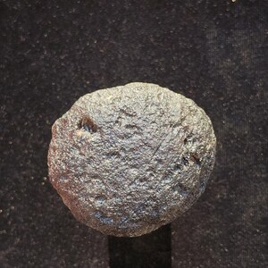 May include: A round, dark grey stone with a rough, textured surface. The object features a pitted, uneven exterior with small indentations and a metallic sheen, displaying a natural, weathered appearance with varied dark grey and charcoal tones.
