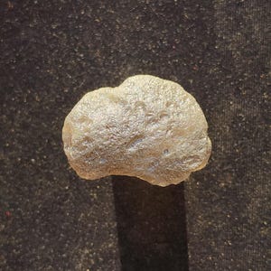 Puede incluir: Una piedra pequeña de forma irregular con una apariencia beige pálida y translúcida. La superficie de la piedra está texturizada con numerosos hoyos, protuberancias y crestas diminutas, creando un aspecto orgánico y rugoso que capta la luz.
