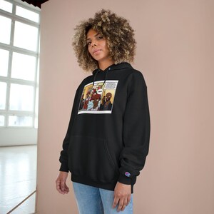 May include: Black Champion hoodie with a comic strip graphic. The graphic shows figures in a scene with speech bubbles. The hoodie has a drawstring hood and a kangaroo pocket. The Champion logo is visible.