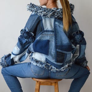 May include: A patchwork denim jacket featuring various shades of blue fabric, frayed edges, and decorative fabric rose embellishments on the shoulders. The jacket has a high collar with a raw, distressed hem and long sleeves with frayed cuffs.