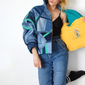 May include: A patchwork denim jacket with green accents and a yellow interior featuring an embroidered cartoon mouse. The jacket is paired with blue jeans. The model is wearing a black top and black socks.
