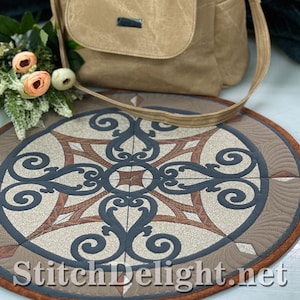 May include: A round, quilted mat with a detailed, symmetrical design in shades of beige, brown, and navy blue. The mat features a central geometric pattern with swirling accents and a decorative border. A tan shoulder bag rests above the mat. The website "StitchDelight.net" is printed on the mat.