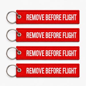 Op de afbeelding: Vier rode rechthoekige sleutelhangers met de witte tekst "REMOVE BEFORE FLIGHT". Elke sleutelhanger heeft een zilveren metalen ring voor bevestiging.