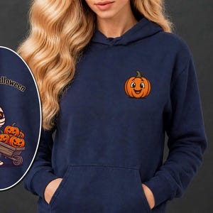 Może przedstawiać: Granatowa bluza z kapturem z małą grafiką pomarańczowej dyni na piersi. Owalna wstawka przedstawia rysunkową mumię pchającą drewnianą taczkę wypełnioną trzema pomarańczowymi dyniami, z napisem Happy Halloween powyżej.