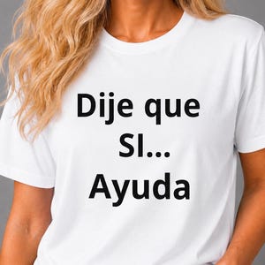 Op de afbeelding: Een wit T-shirt met korte mouwen en zwarte tekst op de voorzijde. De tekst luidt "Dije que SI... Ayuda" in een eenvoudig, vet, schreefloos lettertype. Het kledingstuk is voorzien van een klassieke ronde hals en een standaard pasvorm.