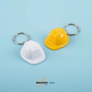 May include: Two miniature hard hat keychains, one white and one yellow, rest on a light blue surface. Each keychain features a silver ring. The keychains are a construction-themed item.