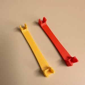 May include: Two elongated, flat, 3D-printed plastic components, one yellow and one red. Each piece features a straight, narrow body with a small, U-shaped bracket or clip mechanism at both ends, showing a textured surface finish from the printing process.