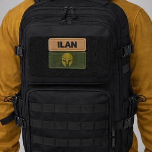 May include: Black tactical backpack with multiple compartments, MOLLE webbing, and adjustable straps. Features a rectangular patch with the name "ILAN" and a green patch with a helmet graphic. The backpack is designed for durability and functionality.