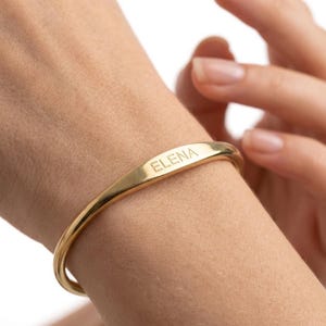 Op de afbeelding: Een goudkleurige armband met de naam "ELENA" gegraveerd op de voorkant. De armband heeft een eenvoudig, gebogen ontwerp en wordt om een pols gedragen. De afbeelding is een close-up van de armband.