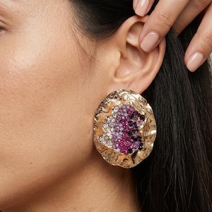 May include: Gold-toned, textured, oval-shaped earring with a cluster of purple, pink, and clear crystals. The earring is worn on the ear, with a hand gently touching the hair. The jewelry has a hammered metal appearance.