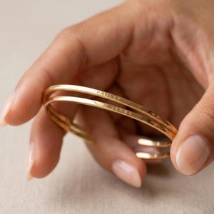 May include: Gold-coloured, stackable bracelets engraved with the names "HAILEY" and "JENNA". The bracelets are thin and have a smooth, polished finish. The bracelets are being held by a person.