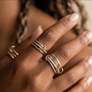 Puede incluir: Primer plano de varios anillos con un diseño plateado y dorado. Los anillos presentan bandas plateadas, detalles dorados y pequeñas piedras brillantes. Los anillos se llevan en los dedos, mostrando sus intrincados detalles y su elegante estilo.