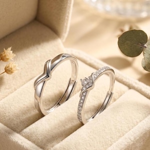 Op de afbeelding: Twee zilveren ringen in een beige ringendoosje. Eén ring heeft een gedraaid ontwerp, de andere heeft een vierkante cluster van kleine, heldere stenen en een band van kleinere stenen. De ringen zijn waarschijnlijk verstelbaar.