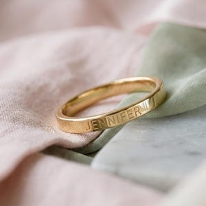 Può includere: Un anello color oro con il nome "JENNIFER" inciso sulla fascia. L'anello è presentato su uno sfondo di tessuto rosa e verde. Il design dell'anello è semplice e moderno. Un gioiello classico.