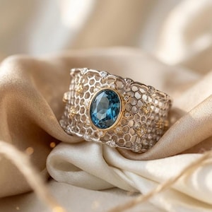 Könnte beinhalten: Ein breiter Silberring mit Wabenmuster, der einen großen ovalen blauen Edelstein in einer Goldfassung aufweist. Kleine Diamanten und goldene Akzente sind um den Ring gesetzt. Der Ring wird auf einem cremefarbenen Satinstoff präsentiert.