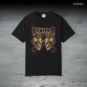 May include: Black t-shirt featuring the word "PANTERA" in a distressed font above two golden skulls with horns. Red starburst designs are behind the text and skulls. The shirt is a classic crew neck style.
