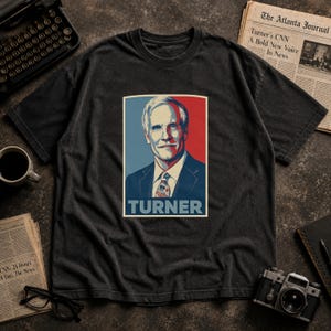 May include: Dark gray t-shirt featuring a portrait of a man with the name "TURNER" below. The portrait is set against a red and blue background. The shirt is surrounded by a typewriter, newspaper, and a camera.