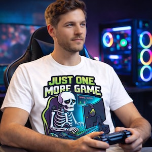 A man wearing a white t-shirt with a graphic of a skeleton playing a video game, with the text "Just one more game" printed on it.