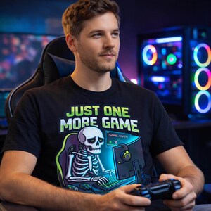 A man wearing a black t-shirt with a graphic of a skeleton playing a video game, holding a game controller, and looking to the side.
