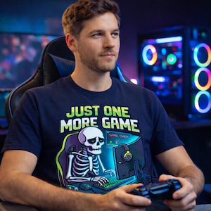 A man wearing a t-shirt with a graphic of a skeleton playing a video game, sitting in a gaming chair in front of a computer monitor.