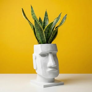May include: A white Moai head-shaped planter, featuring a snake plant with green and yellow striped leaves. The planter is on a white surface, set against a vibrant yellow backdrop.