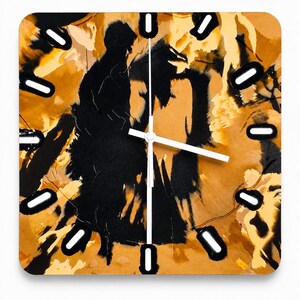 May include: A square wall clock featuring an abstract design with bold black ink-like shapes against a warm ochre and tan background. The clock face has twelve white rectangular hour markers and three thin white hands centered on the dial.