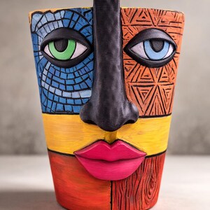 Może przedstawiać: Kolorowa ceramiczna donica w kształcie twarzy z czarnym nosem i oczami z zielonymi i niebieskimi tęczówkami. Twarz podzielona jest na sekcje z niebieskiej mozaiki, pomarańczowych geometrycznych wzorów, żółtego, czerwonego i tekstur słojów drewna. Usta są w kolorze jasnoróżowym.