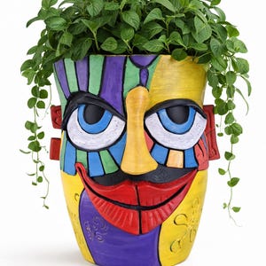 May include: A colorful, hand-painted ceramic planter shaped like a face. The planter features a yellow background with a face design in red, blue, purple, green, and black. Green plants spill over the top of the planter.