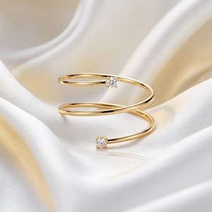 Könnte beinhalten: Ein goldfarbener Spiralring mit zwei kleinen, klaren Edelsteinen. Der Ring ist vor einem weißen, seidigen Hintergrund platziert, was eine luxuriöse und elegante Ästhetik erzeugt. Die Edelsteine sind rund und funkeln.