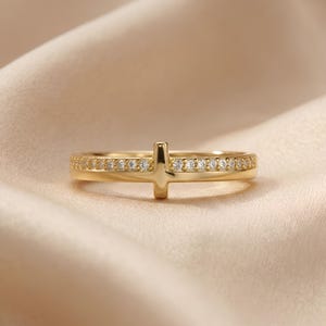 Puede incluir: Un anillo dorado con diseño de cruz. El anillo presenta una banda con pequeñas piedras preciosas transparentes. La cruz está centrada en el anillo y también es dorada. El anillo se encuentra sobre un fondo suave y de tono neutro.