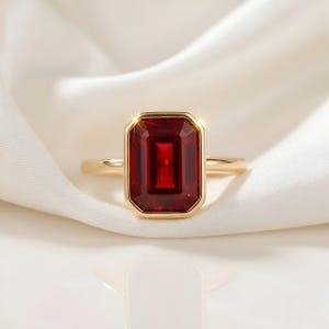May include: A gold ring featuring a large, rectangular, deep red gemstone. The gemstone is set in a gold bezel, and the ring is displayed on a white, silky fabric. The ring's band is a simple, polished gold.