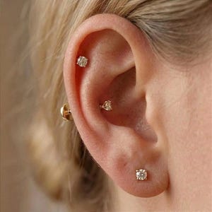 May include: Three gold stud earrings with clear round-cut stones are worn in the ear. One earring is placed in the lobe, one in the conch, and one in the upper cartilage. Each piece features a metallic gold setting holding a single sparkling stone.